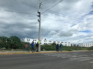 Personas esperando el camión sobre el borde de una calle en Tlajomulco, a lo lejos se ven edificios y arriba el cielo nublado