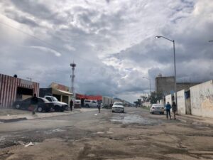 Calle descuidada de Tlajomulco con carros en las laterales, dos personas caminando y el cielo nublado arriba