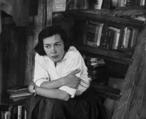 Mujer adulta con brazos cruzados blusa blanca y detrás estante de libros. Fotograma de Loving Highsmith