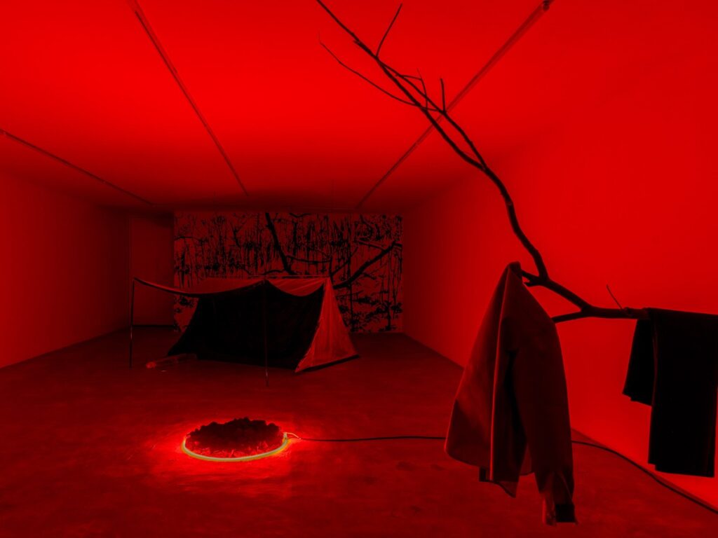 Instalación de arte con una casa de campaña, fogata y prenda colgada en rama en habitación roja