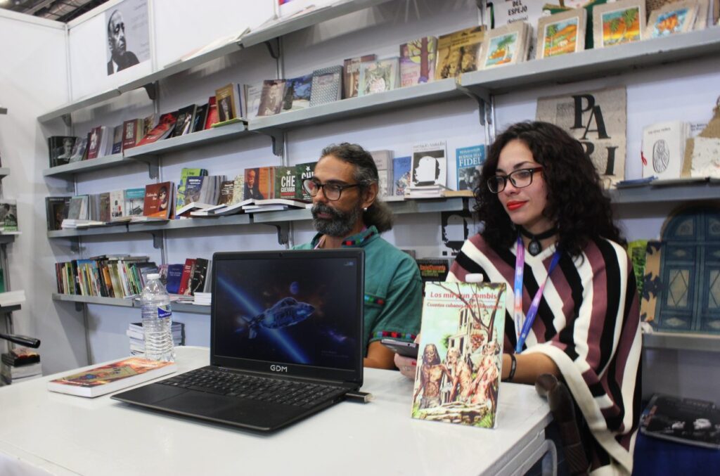 Hombre adulto con barba y lentes y mujer joven cabello negro con lentes sentados escritorio con computadora encima