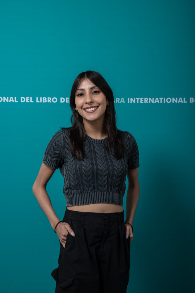 Mujer sonriendo cabello negro suéter gris manos en pantalones Larissa Rú