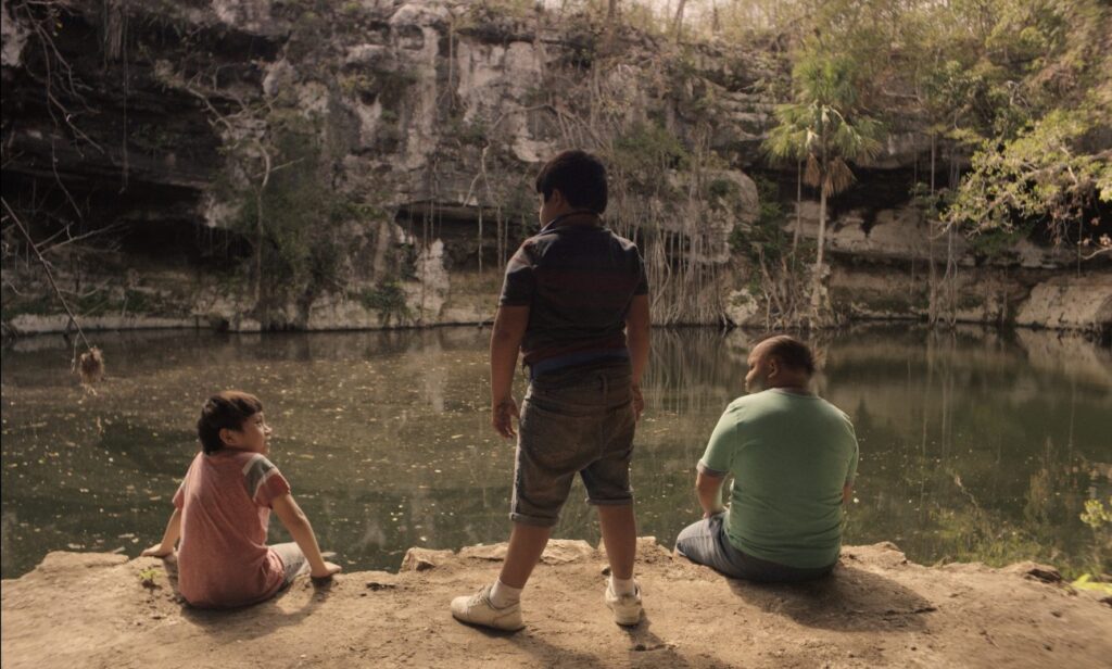 Tres personas sentadas al borde de un cenote. Niño de pie. Monstruo de Xibalba.