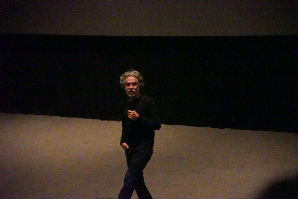 Hombre adulto vestido de negro con microfono en la mano en sala oscura